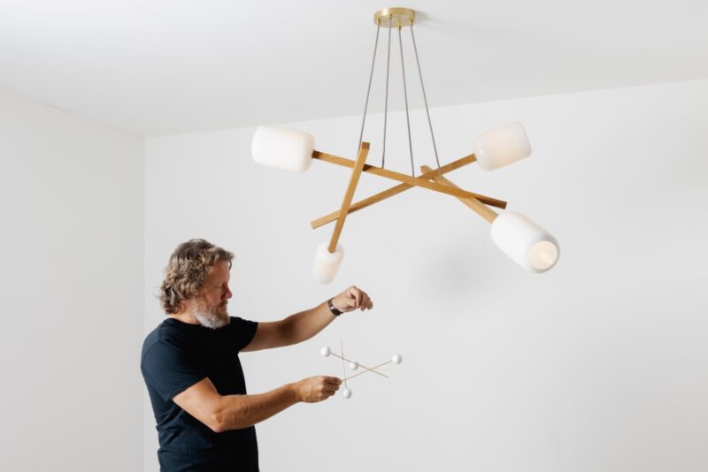 A man holding a small model of a modern chandelier—reminiscent of a Nick Sheridan Cerno design—stands under a large, similarly styled chandelier in a pristine white room.