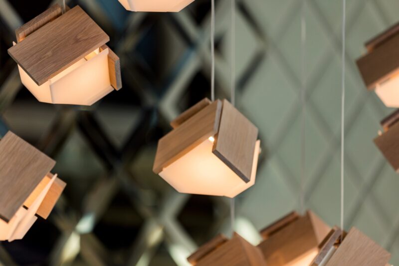 Several modern pendant lights with wooden geometric shades, inspired by Nick Sheridan Cerno designs, hang from the ceiling, with a blurred background of a lattice-patterned wall.