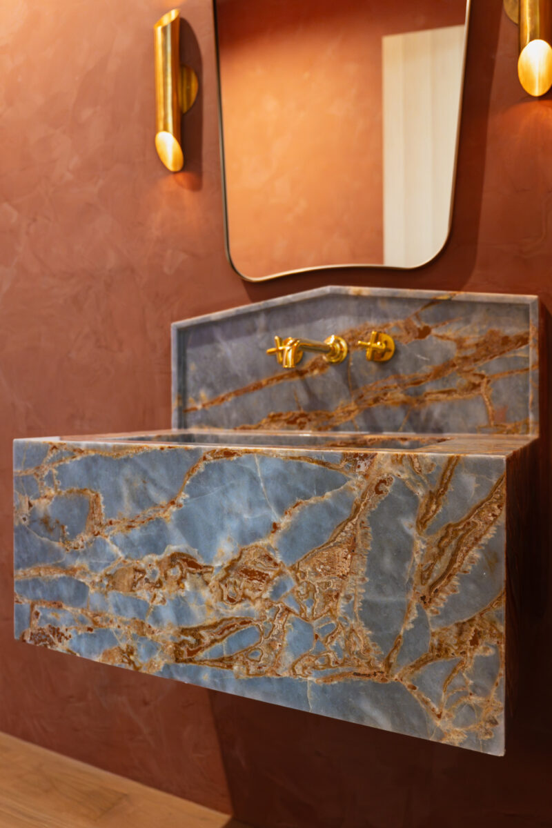 Blue and brown marble sink from April Graves Aria Stone Gallery with a brass wall-mounted faucet, rectangular mirror, and brown textured wall background.