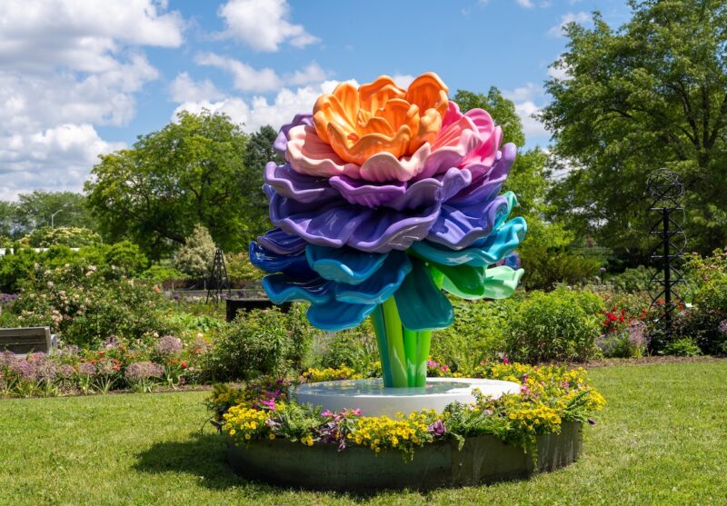 Large, multicolored abstract flower sculpture stands on a circular platform surrounded by yellow flowers in a garden under a partly cloudy sky.