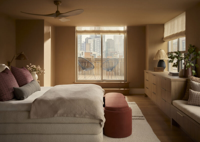Bedroom with beige walls, bed with pillows, red bench, wooden dresser, large window, city buildings view, and two chairs on the balcony.