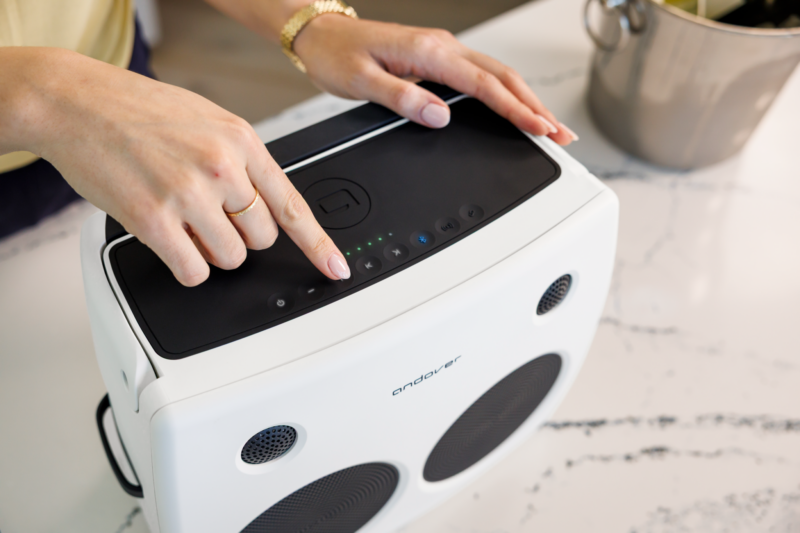 A person presses a button on top of a white Andover Audio FreePlay device with speakers, placed on a white countertop near a stainless steel pot.