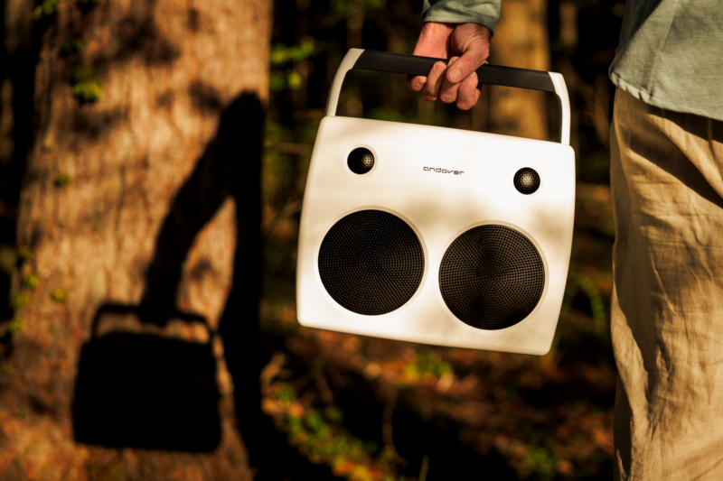 A person is holding an Andover Audio FreePlay, a sleek white portable speaker with a handle, outdoors near trees, casting a shadow onto a tree trunk.