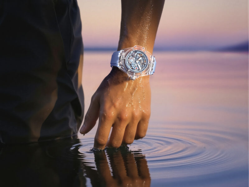 A person’s wet hand wearing a transparent Hublot Big Bang Sapphire Sky Blue wristwatch is partially submerged in calm water at sunset, creating gentle ripples on the surface.