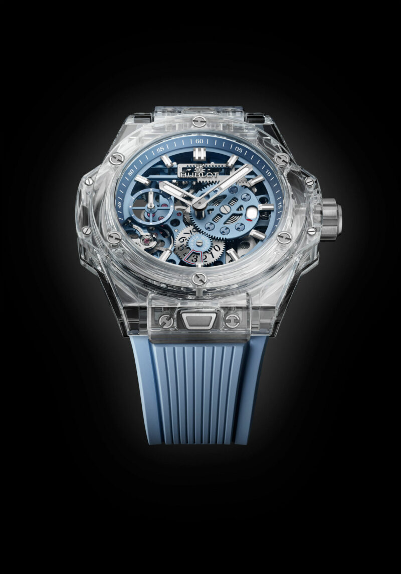 The Hublot Big Bang Sapphire Sky Blue transparent wristwatch features visible blue and silver inner mechanisms, a striking blue rubber strap, and a clear casing, all set against a black background.