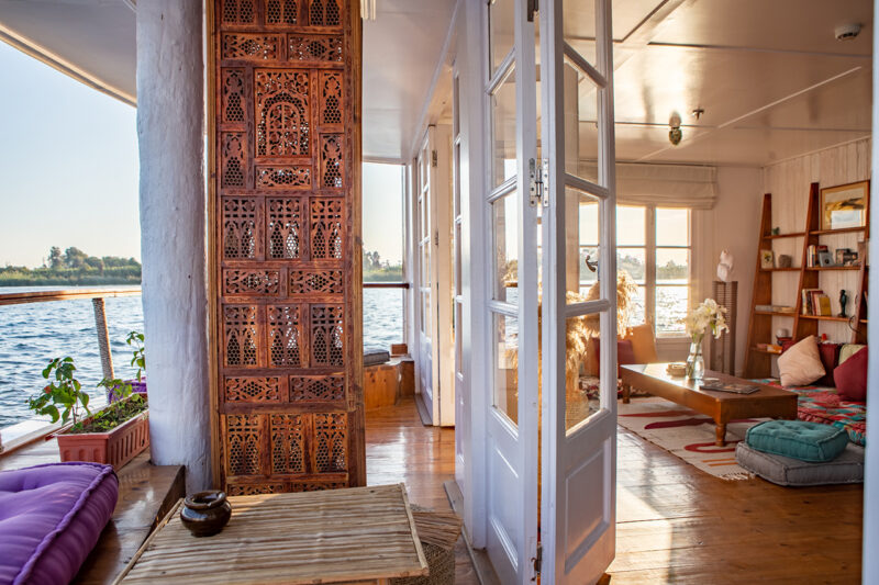 Sunlit houseboat interior with wooden floors, large windows, ornate carved panel, cushions, low table, and plants. Water and shoreline visible through windows and open doors.