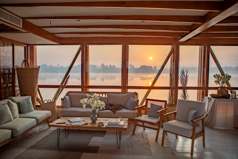 A cozy lounge with modern wooden furniture overlooks a calm lake at sunrise through large windows, creating a peaceful atmosphere.