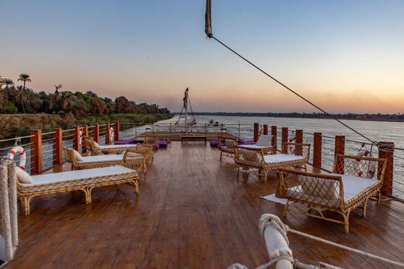 Open deck of a riverboat with wicker lounge chairs facing the water, wooden flooring, and railings, set against a calm river and a sunset sky.