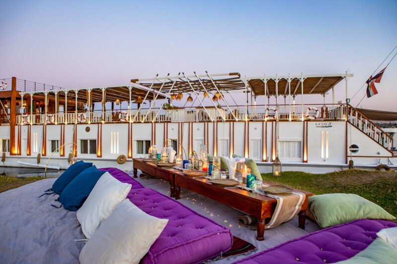Outdoor picnic setup with colorful cushions and a low wooden table arranged on a lawn next to a docked riverboat at sunset.