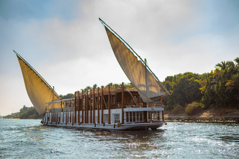 A large traditional sailboat with two triangular sails moves along a river, with green trees lining the shore in the background under a partly cloudy sky.