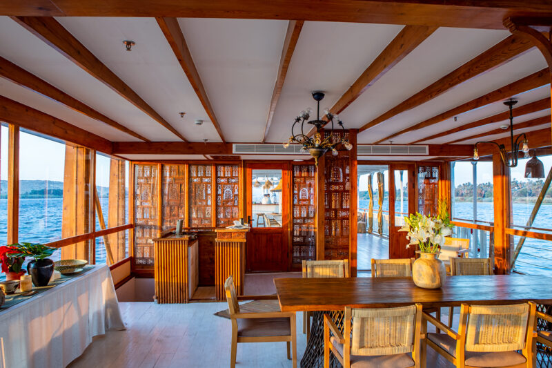 Interior of a wooden boat with large windows, a dining table with chairs, decorative flowers, and views of water and distant land through the windows.