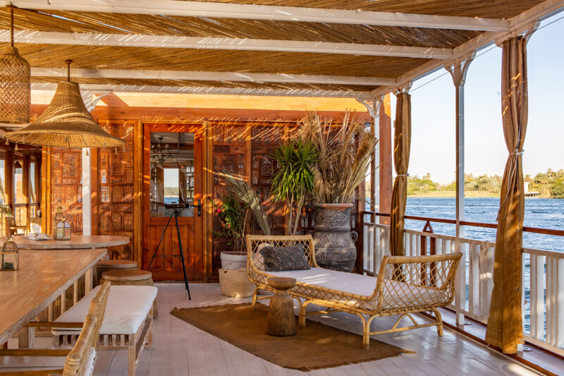 Sunlit boat deck with wicker furniture, potted plants, wooden decor, and river view; shaded by a bamboo roof.