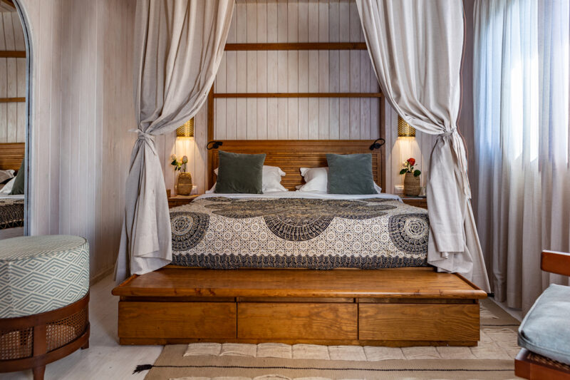 A neatly made bed with patterned bedding, green pillows, and beige curtains on a wooden frame, set in a bright bedroom with light walls and soft natural light from a window.