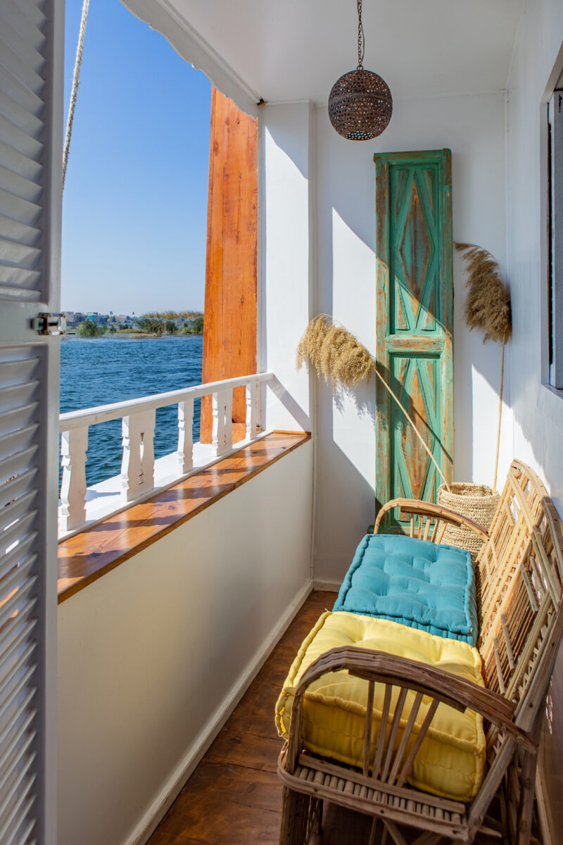 A narrow balcony with wicker chairs, colorful cushions, a wooden railing, dried grass decor, a green rustic door, and a view of water under a clear blue sky.