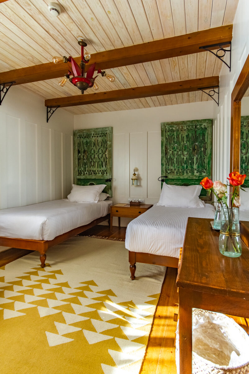 A bedroom with two single beds, a wooden ceiling, green wall art, a yellow patterned rug, and a wooden dresser with vases of red and orange flowers.