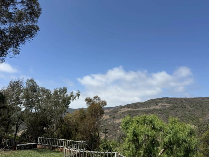 A clear sky with scattered clouds above green hills and trees, with a white fence in the foreground.