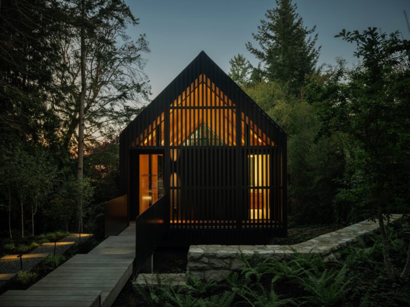 Modern black wood cabin with slatted exterior and warm interior lighting, the Little Giant forest studio is surrounded by trees and greenery at dusk, viewed from a raised walkway.