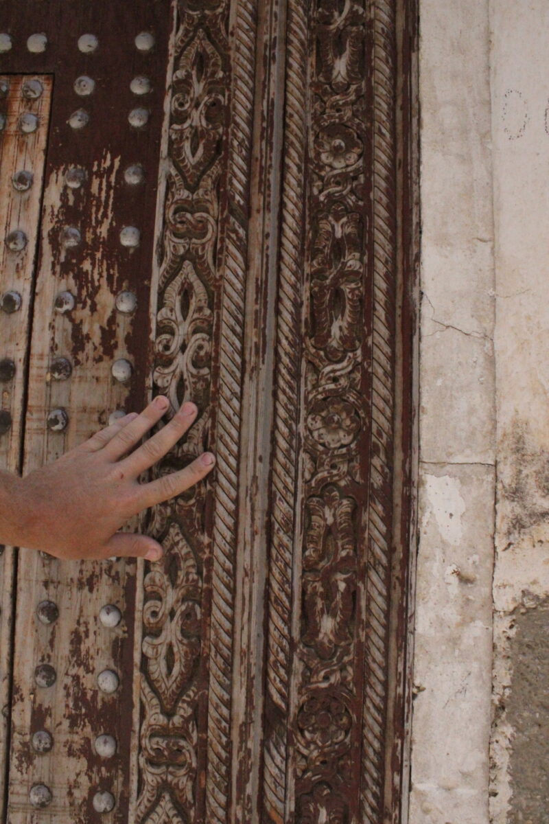 A hand touches an ornate, weathered wooden door with intricate carvings, metal studs, and a stone wall on the right side.