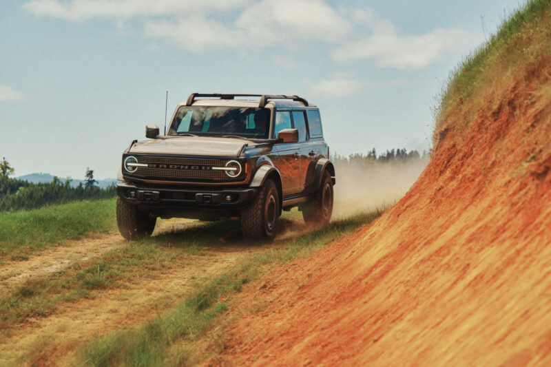 A Ford Bronco drives on a dirt trail next to a steep, orange-colored hillside under a partly cloudy sky.