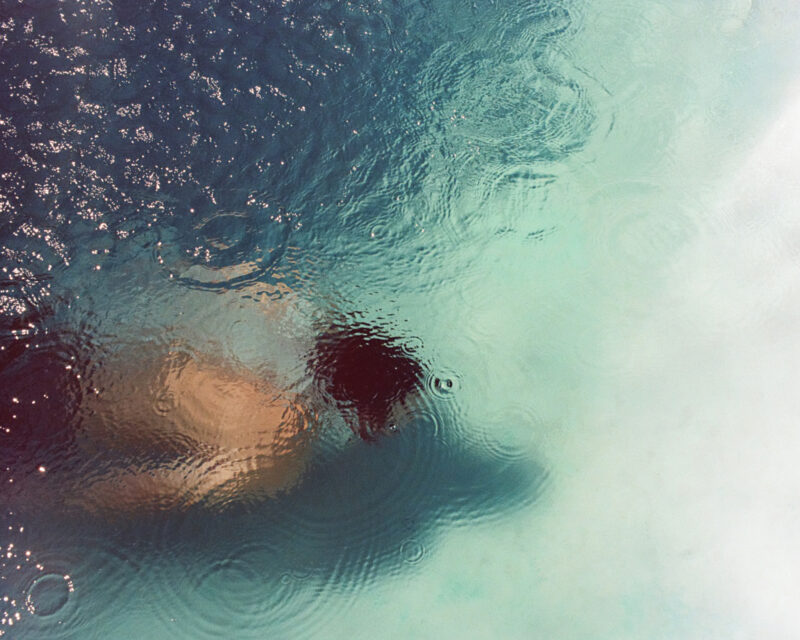Person swimming underwater with ripples and raindrops visible on the surface of the water.