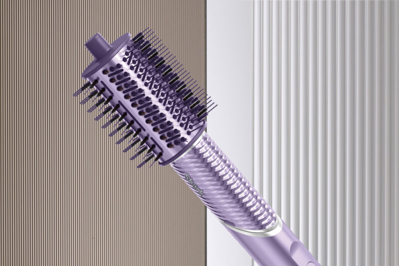 A close-up of a purple rotating hot air brush for hair styling, reminiscent of the Shark FlexStyle IonCurl, featuring bristles and a textured handle, set against a striped background.