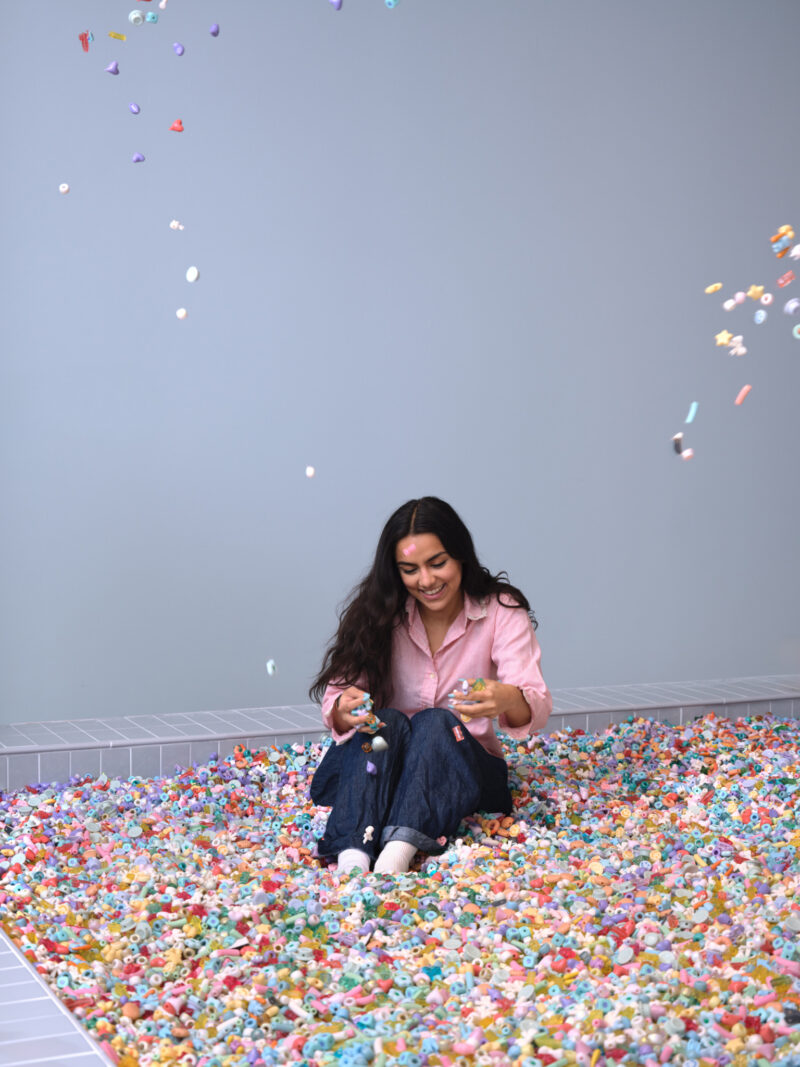 A woman sits on a floor covered with colorful small toys, smiling and holding toys in her hands while more toys fall around her against a plain light blue wall, evoking the playful atmosphere of a Cj Hendry Pastel exhibition.