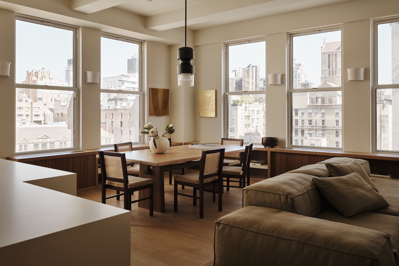 Modern dining and living area with large windows, wooden table and chairs, beige sofa, pendant light, and cityscape view outside.