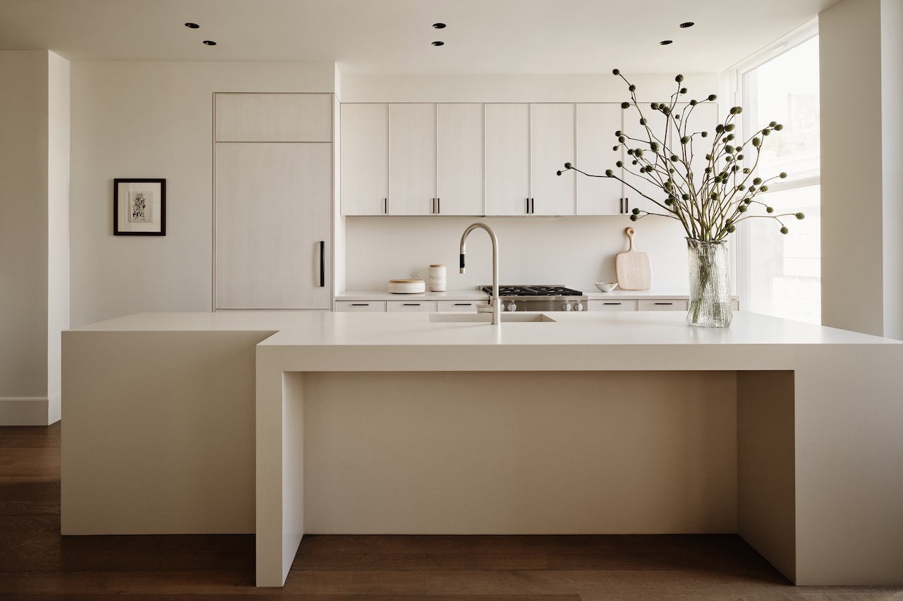 Minimalist kitchen with white cabinets, a large island with a built-in sink, wooden floors, and a glass vase with tall branches on the counter.