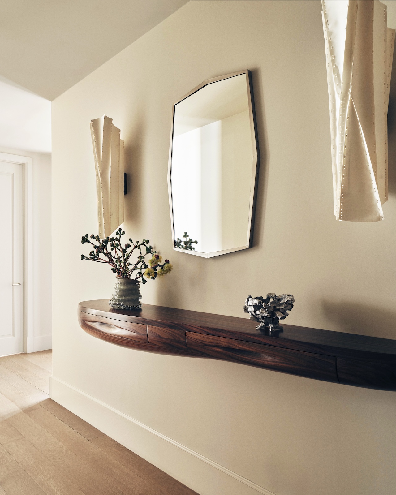 A hallway with a wooden floating console, a vase with branches, a decorative bowl, an octagonal wall mirror, and two sculptural wall sconces.