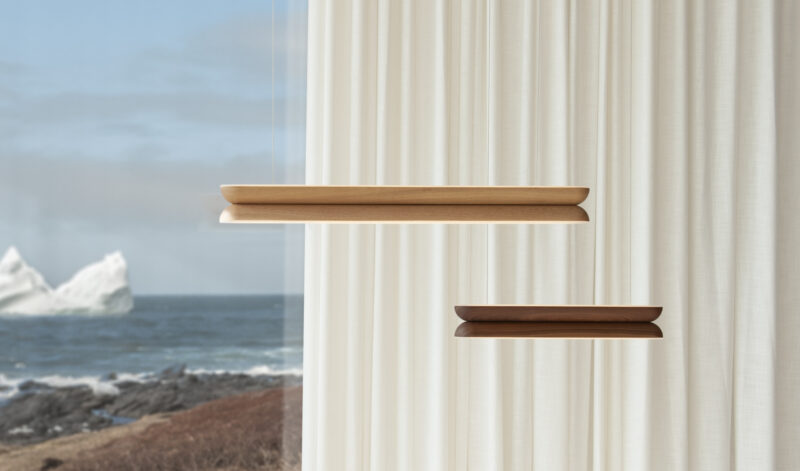Two wooden pendant lights from the Beaufort lighting collection hang in front of sheer white curtains, with a coastal landscape featuring rocks, ocean, and icebergs visible through a window in the background.