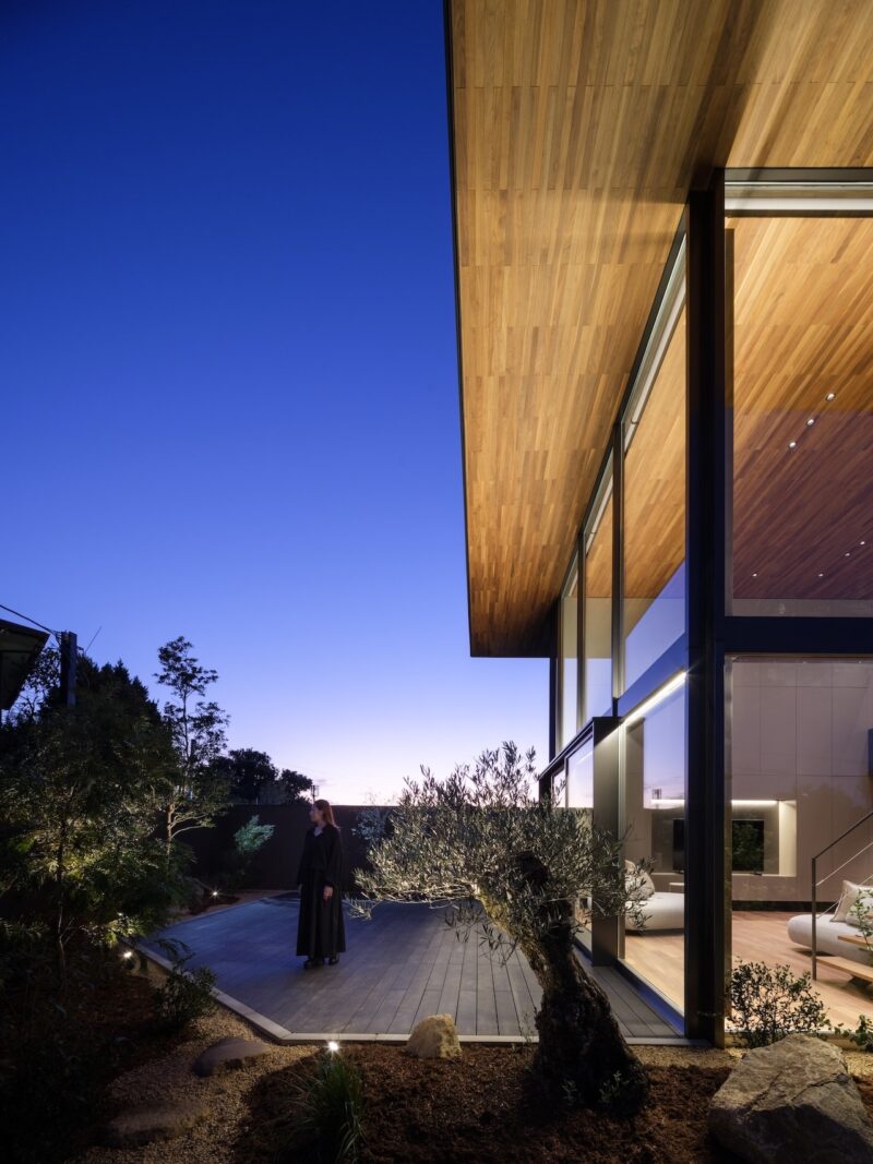 A person stands on a wooden deck outside a modern house with large glass windows at dusk, surrounded by trees and outdoor lighting.
