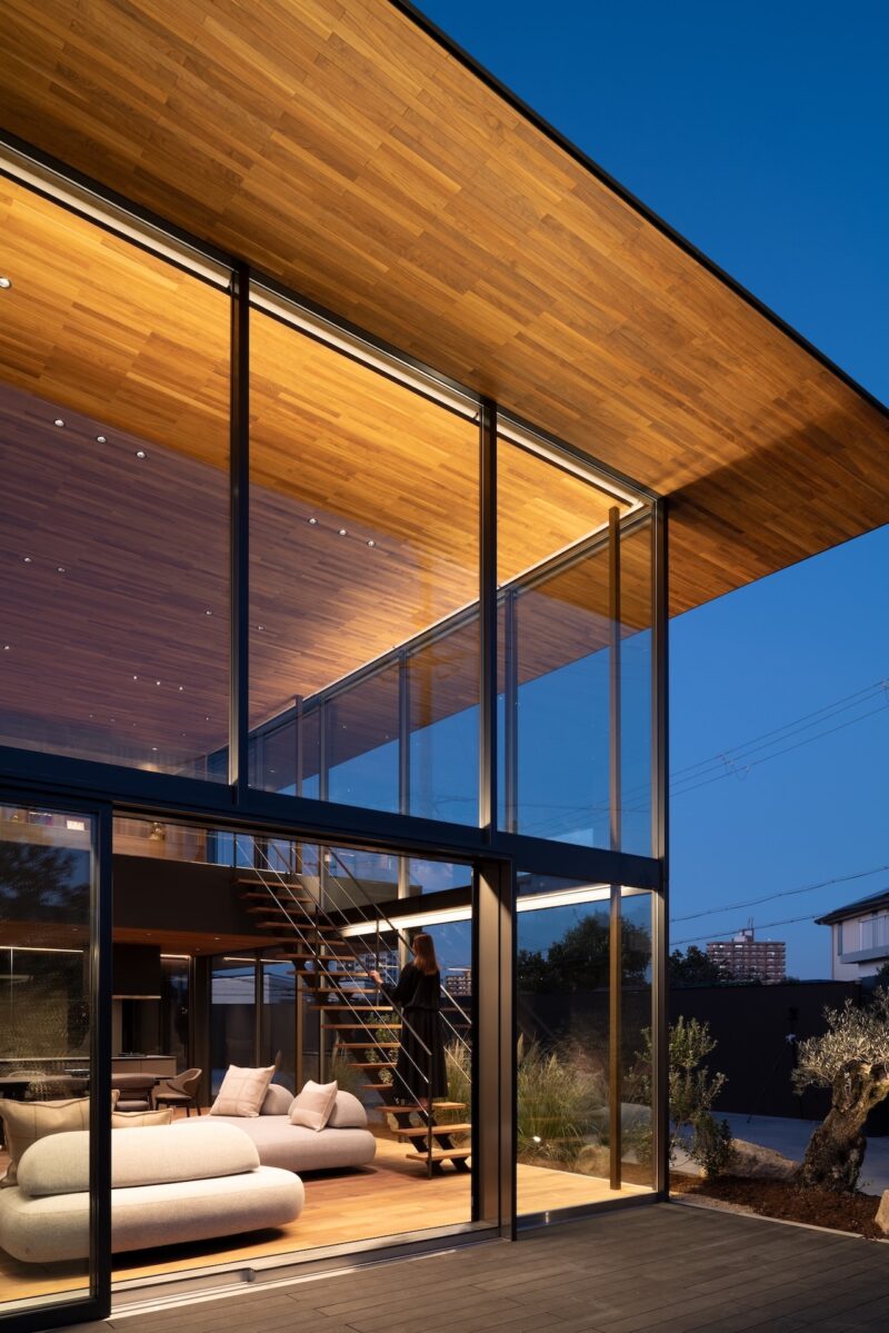 Modern house with large glass walls, wood ceiling, open living area, and staircase visible at dusk. Indoor and outdoor spaces are illuminated.
