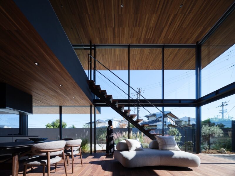 A modern living room with wood floors and ceiling, large glass windows, a floating staircase, and a person standing near the glass wall.
