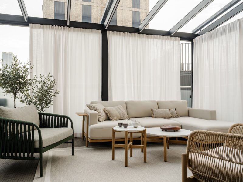 Modern sunroom with large windows, light-colored sectional sofa, rattan armchair, small tables, potted plant, and sheer white curtains. Urban buildings visible outside.