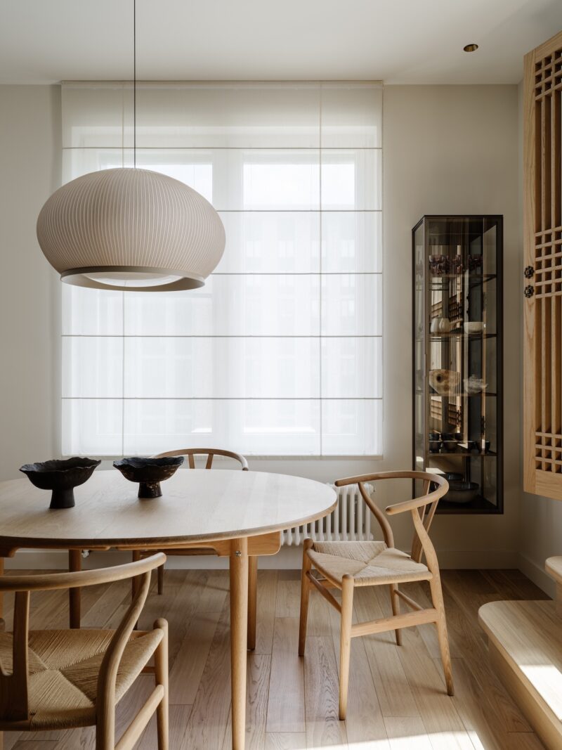 Minimalist dining area with a round wooden table, three wooden chairs, two ceramic bowls, a large hanging lamp, and a glass display cabinet by a window with sheer curtains.
