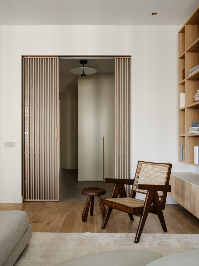 Minimalist living space with a wooden armchair, small round stool, built-in shelving, and slatted sliding doors opening to a hallway.