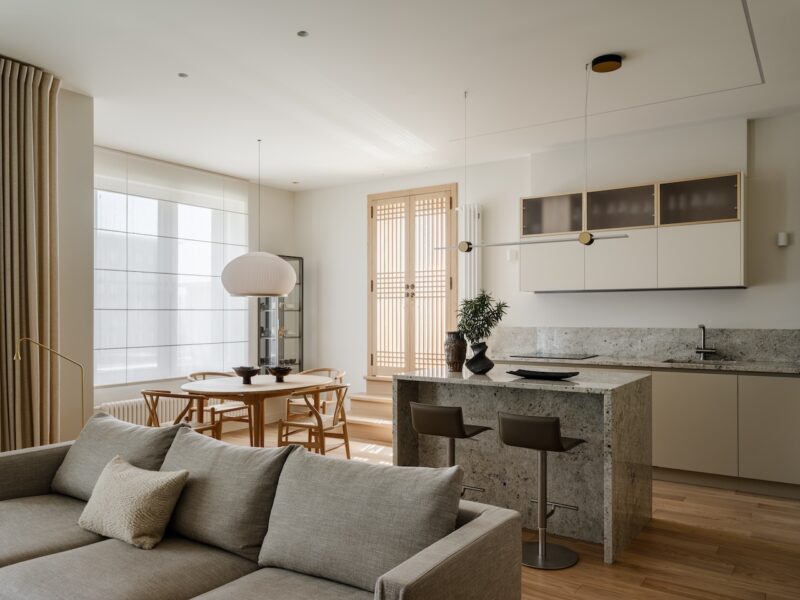 Modern open-plan living space with a gray sofa, dining table, and kitchen with stone island, bar stools, and minimalist decor. Light wood flooring and large windows provide natural light.