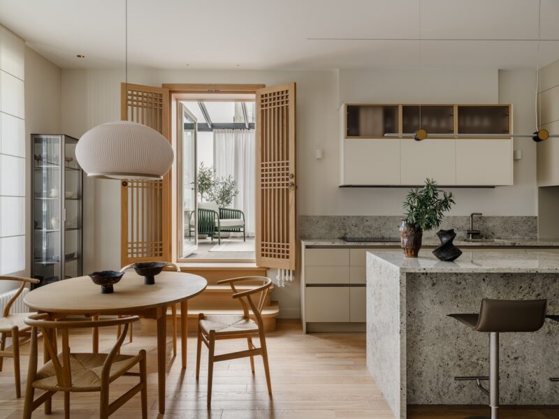 Modern kitchen and dining area with wooden furniture, neutral tones, a round table, island, and open wooden doors leading to a bright room with a green chair and plants.