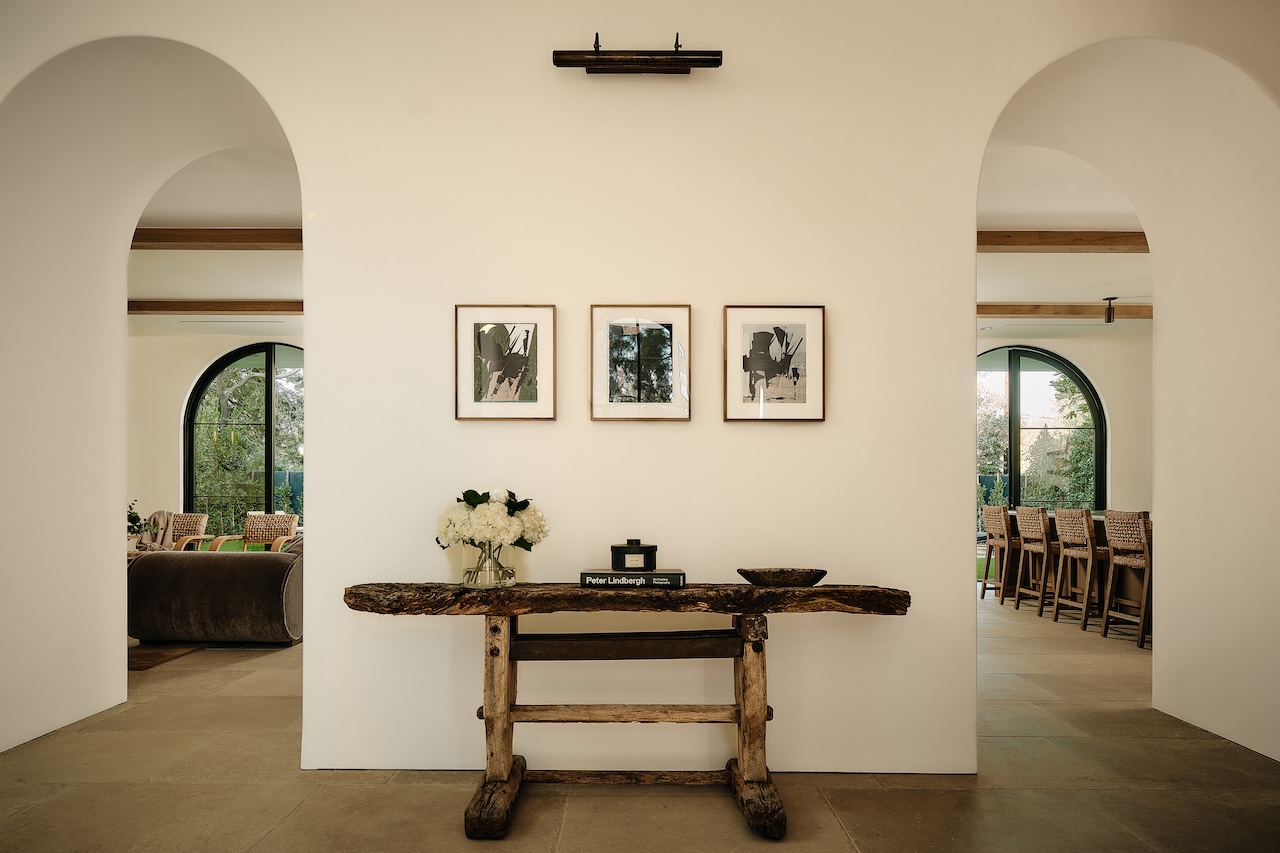 A rustic wooden console table with books and flowers stands against a white wall, beneath three framed artworks; arched doorways reveal a living and dining area.