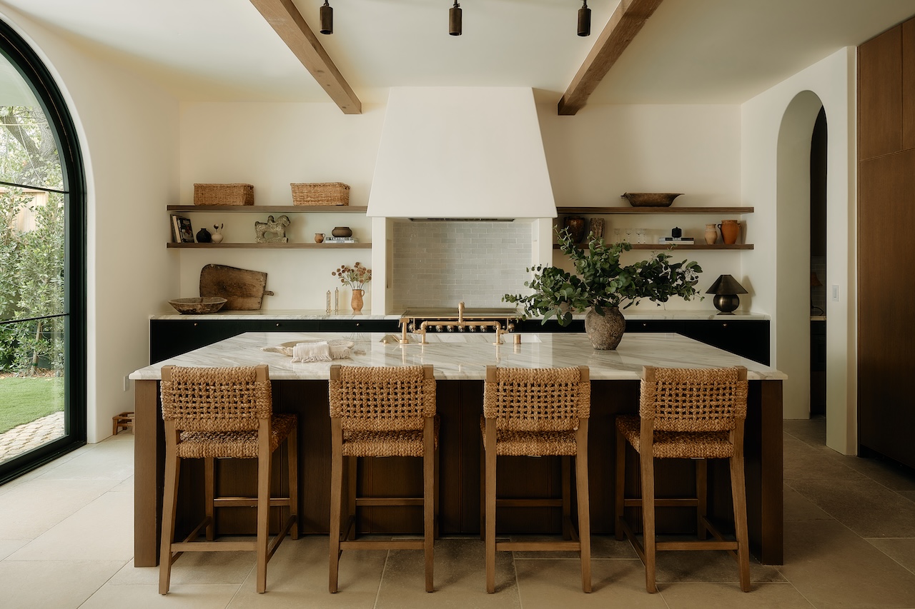 Modern kitchen with a marble island, four wicker barstools, open shelves with decor, wooden beams, and large arched window.