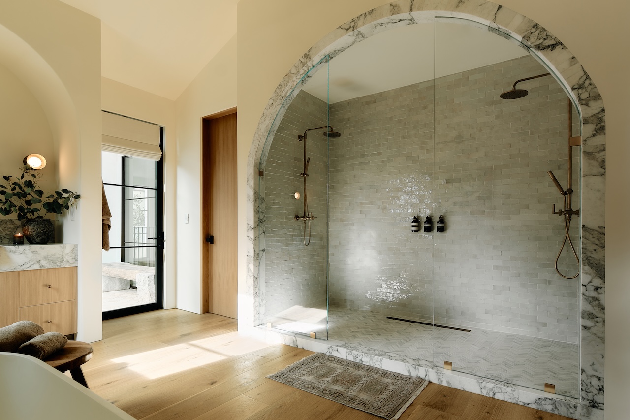 Spacious bathroom with a large, arched glass shower, marble accents, light wood flooring, and a vanity with a potted plant on the left. Natural light enters through nearby windows.