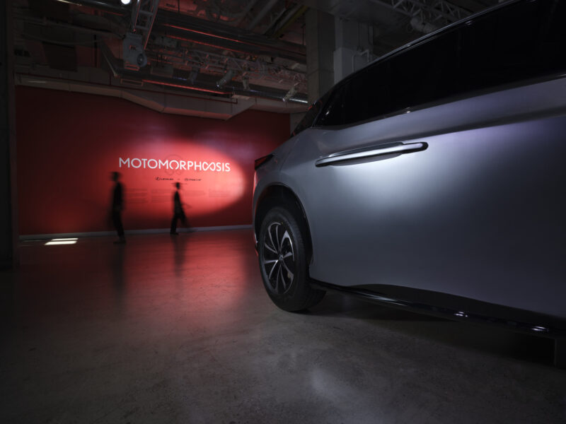 A silver MSCHF Lexus is parked indoors near a red wall displaying the word “MOTOMORPHOSIS” as two people walk by in the background.