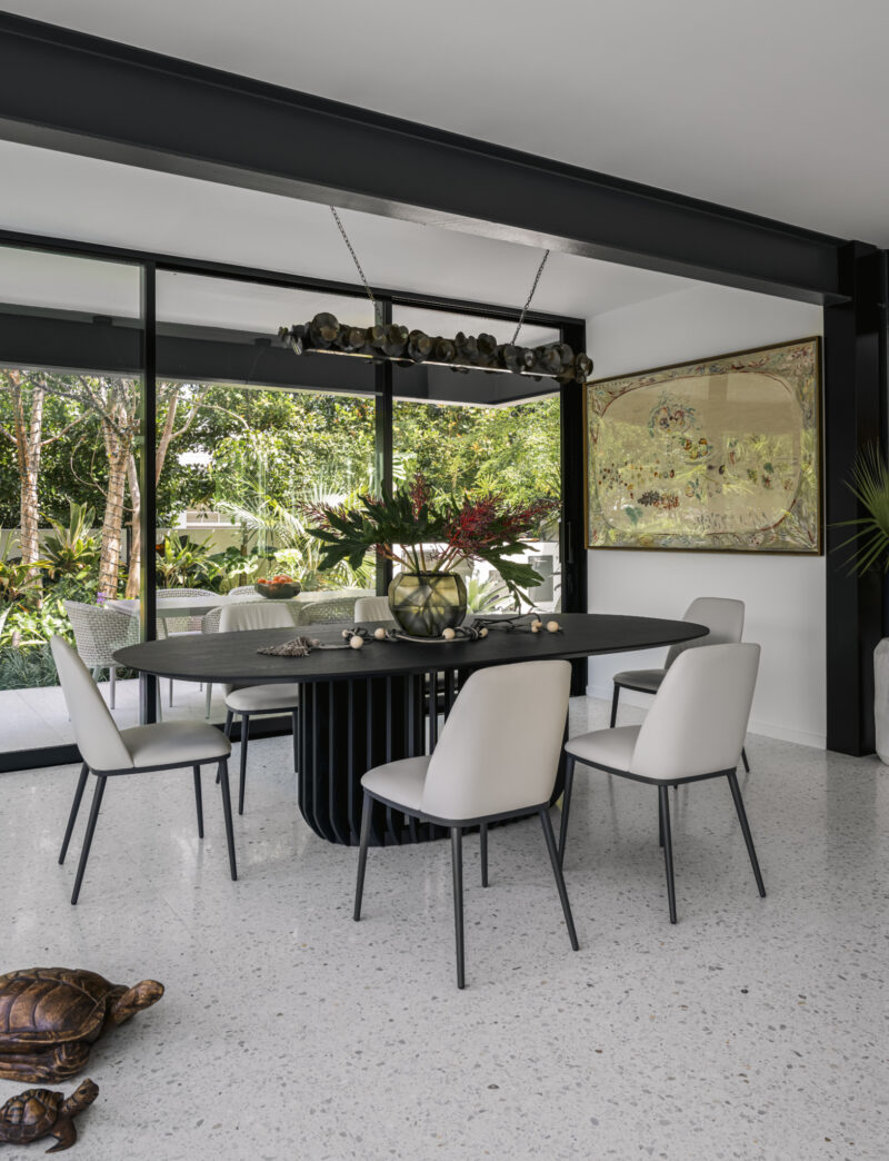 Modern dining room inspired by the Paul Rudolph Biggs Residence, with a black table, six white chairs, wall art, indoor plants, and tortoise shell decor.