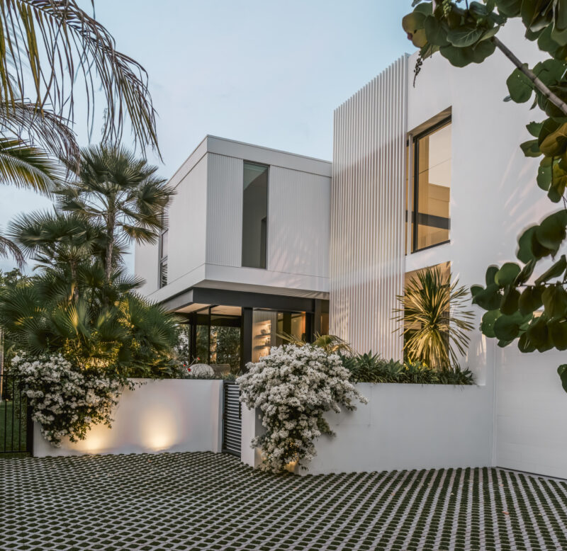 Modern white two-story house with large windows and vertical slats, inspired by the Paul Rudolph Biggs Residence, surrounded by palm trees and flowering shrubs at dusk.