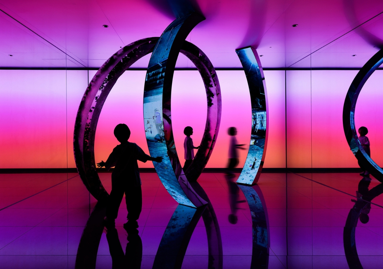 Silhouettes of children interact with large metal rings in a colorful, illuminated room with purple and orange gradient lighting—an innovative space reminiscent of installations recognized by the iF DESIGN AWARD.