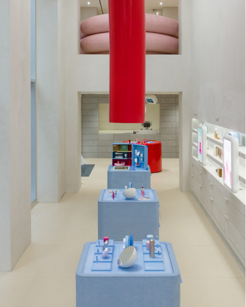 A modern retail store interior, inspired by the Laneige Seoul flagship, with pastel blue display tables, pink circular ceiling accents, a large red ceiling fixture, and white walls with shelves holding products.