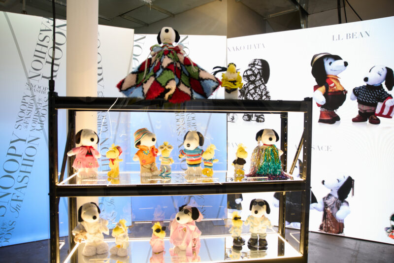 Plush Snoopy and Woodstock dolls dressed in various outfits are displayed on glass shelves, with large illuminated fashion-themed Snoopy images in the background, capturing the playful spirit of Snoopy in Style NYC.