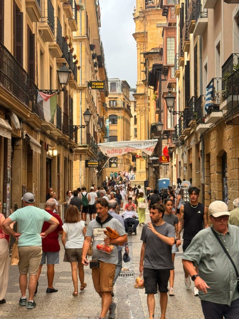 A busy street with many people walking among tall buildings, some with balconies. A banner hangs above the crowd and flags are visible on buildings.