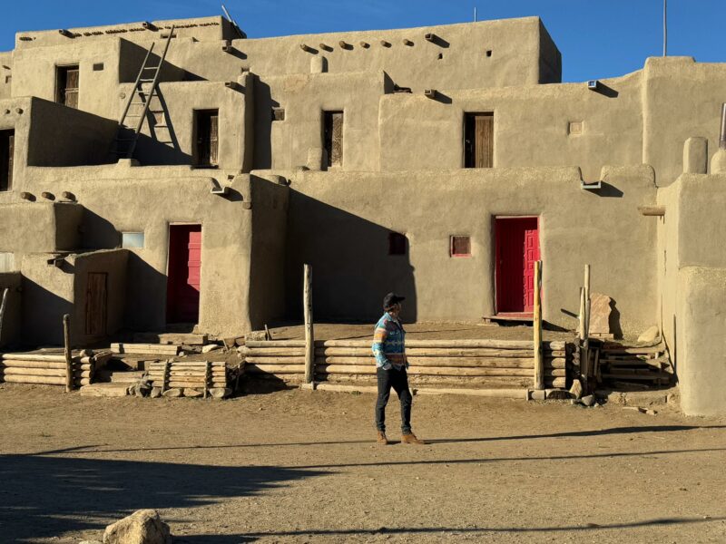 A person walks in front of an adobe building with red doors, wooden ladders, and log railings under a clear blue sky.