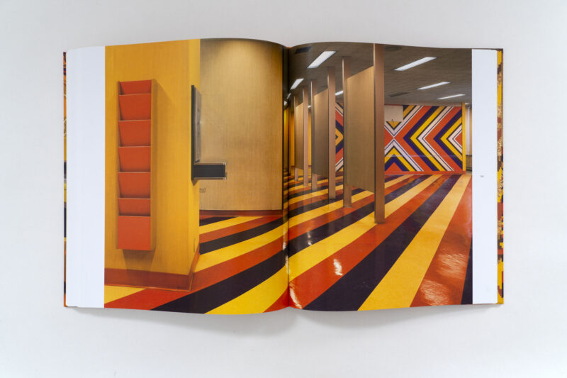An open book showing an interior space with a floor and wall featuring 820 Ebony/Jet diagonal red, yellow, and black stripes, and a vertical orange wall-mounted rack.